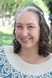 Headshot of a smiling woman with brown, curly hair and green, vegetal background
