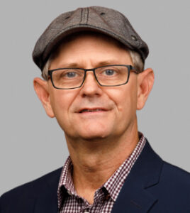 Headshot of a man in glasses and a hat in a suit coat.