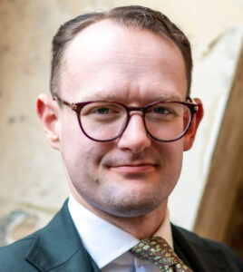 Headshot of a man in glasses and grey suit and patterned tie.
