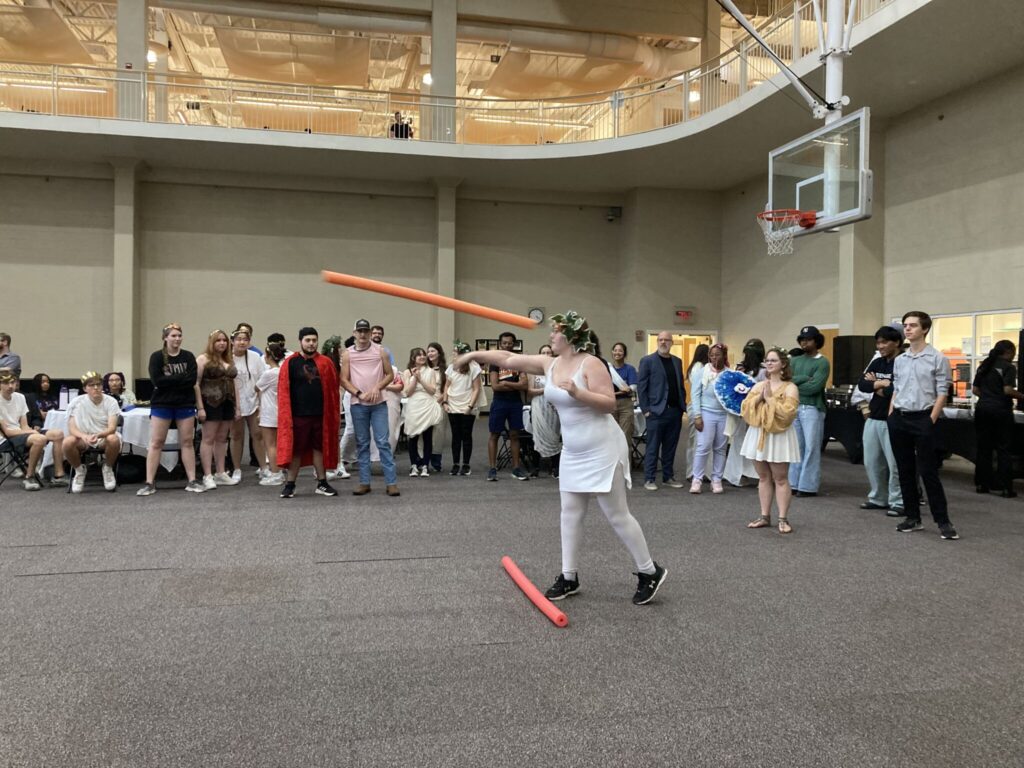 A student throws an orange pool noodle surrounded by other students watching.